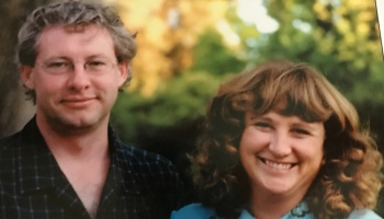 A man with glasses and a smiling woman with curly hair standing together outdoors.