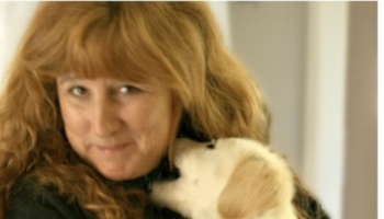 A smiling woman with wavy hair holding a small, light-colored puppy close to her face.