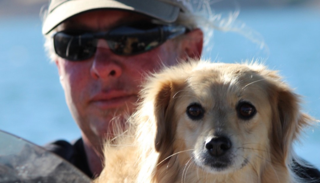 A small, fluffy tan dog looking forward, with a man wearing sunglasses and a cap behind it outdoors by blue water.