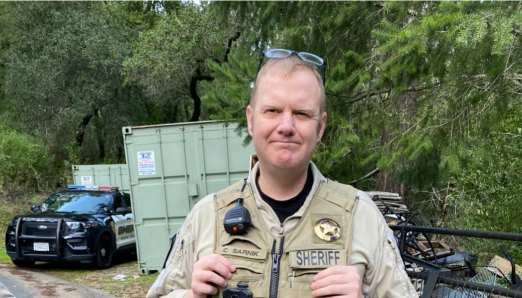 Sheriff's deputy C. Sarnik standing outdoors in uniform with a police vehicle in the background.