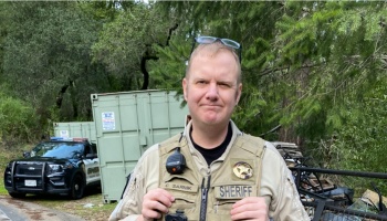 Sheriff's deputy C. Sarnik standing outdoors in uniform with a police vehicle in the background.