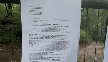 Notice of Trustee's Sale legal document posted on a wire fence in front of a wooded property.