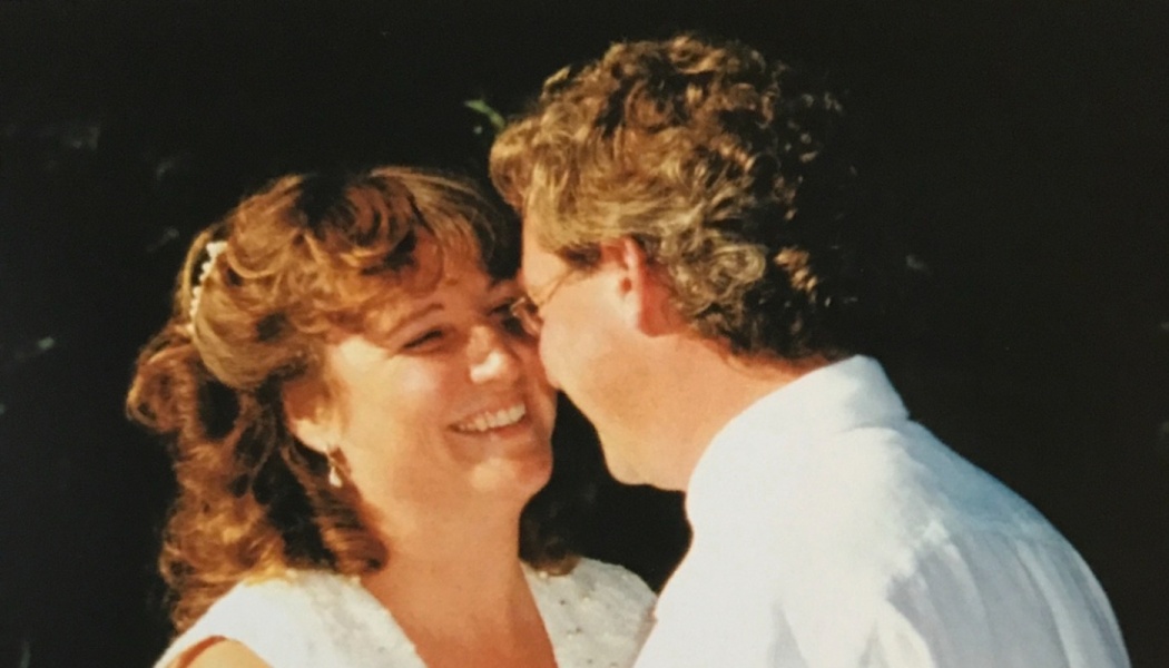 A smiling woman with curly brown hair shares a close, affectionate moment with a man in a white dress shirt.