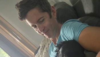 A man in a blue t-shirt smiles gently down at a small black dog resting against him on a couch.