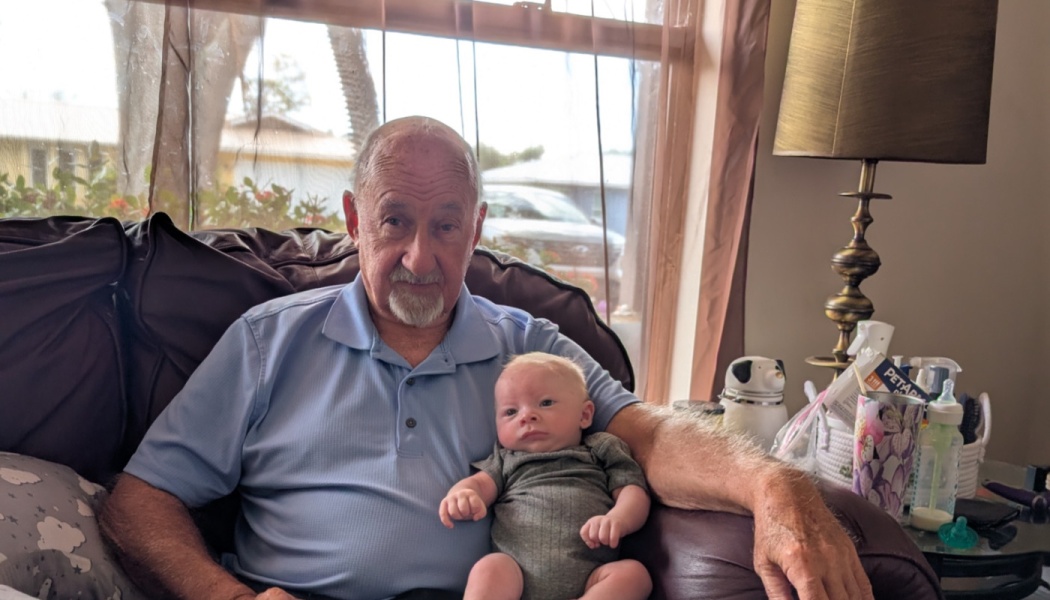 An elderly man in a blue polo shirt sitting in a brown armchair holding a baby on his lap.