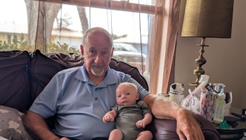 An elderly man in a blue polo shirt sitting in a brown armchair holding a baby on his lap.