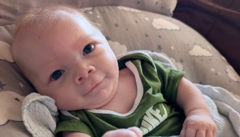 A smiling baby in a green shirt lies on a cloud-and-star patterned pillow, looking into the camera.