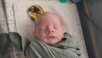 A newborn baby swaddled in a sage green blanket lies sleeping peacefully with eyes closed.