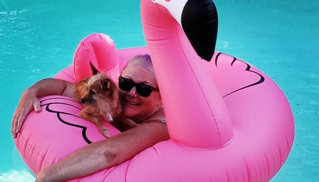 A smiling woman in sunglasses floats in a pool on a giant inflatable pink flamingo while holding her small brown dog.