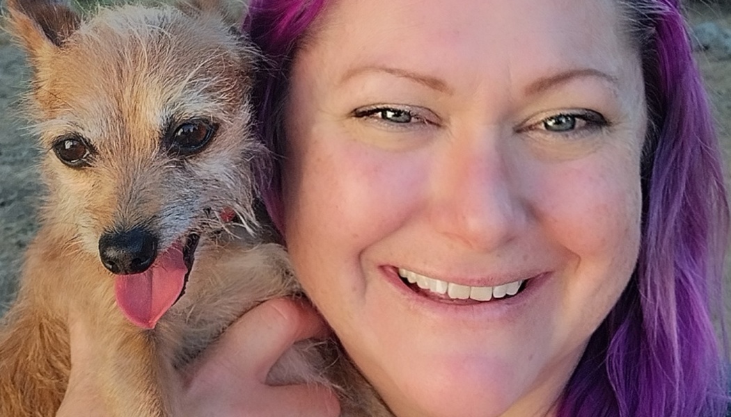 A smiling woman with bright purple hair holds a small, scruffy tan dog with its tongue sticking out.