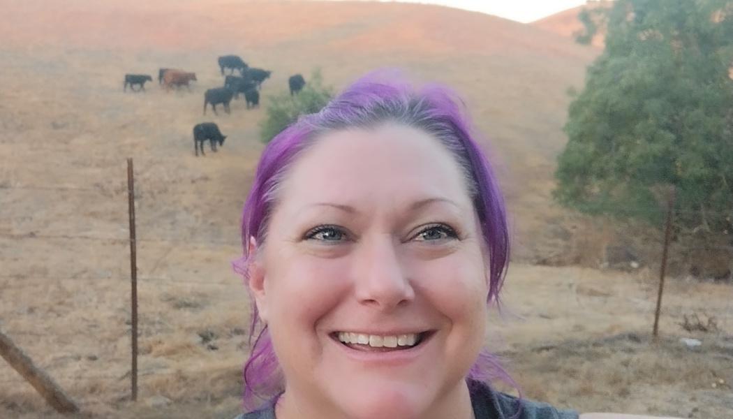 Smiling woman with vibrant purple hair, with a herd of cattle grazing on dry hills behind her.
