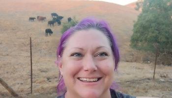Smiling woman with vibrant purple hair, with a herd of cattle grazing on dry hills behind her.