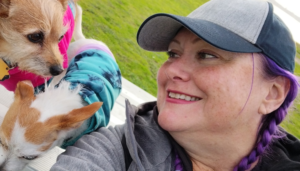 Smiling woman with purple hair taking a selfie outdoors with her two small dogs in colorful clothes.