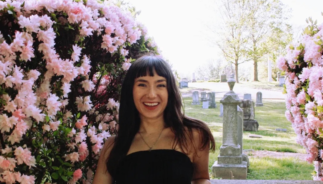 A smiling woman with dark hair framed by blooming pink flowers, with a cemetery in the sunny background.