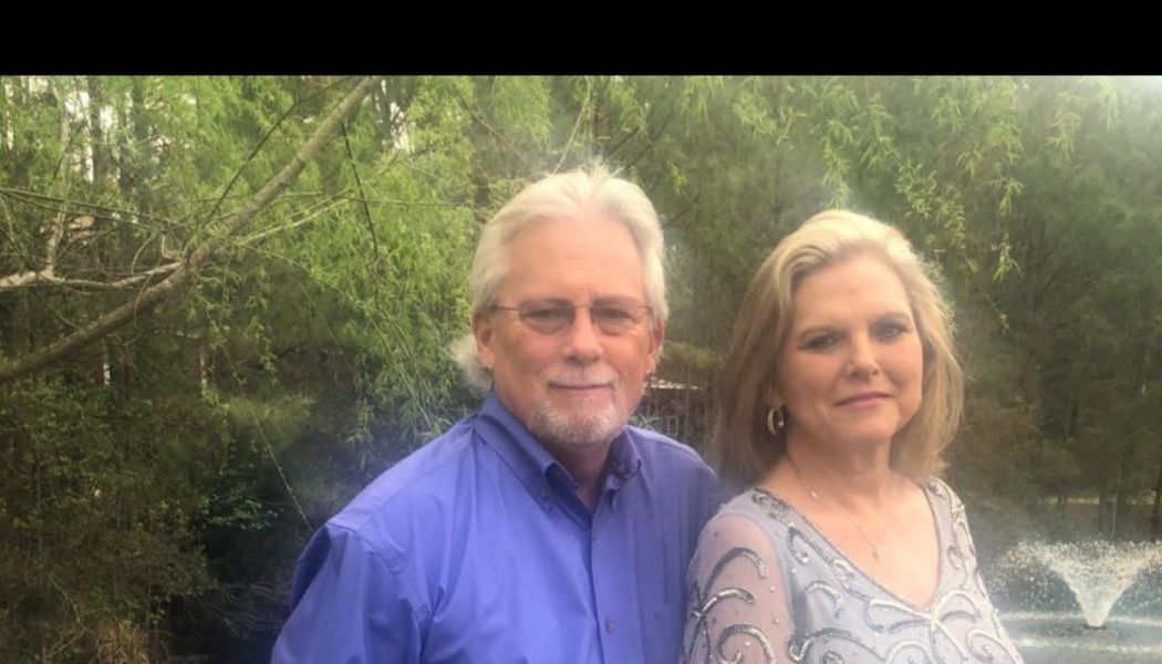 A smiling senior couple standing together outdoors in front of lush green trees and a water fountain.