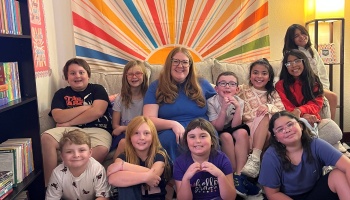 A smiling teacher and her diverse group of happy students sitting together in a colorful classroom reading area.