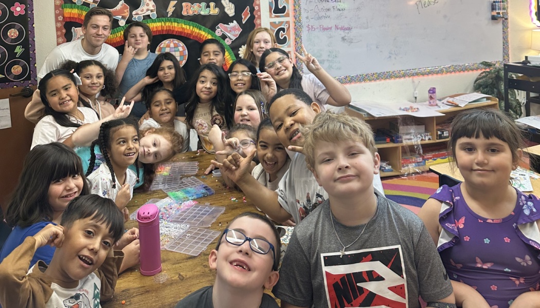A cheerful group of elementary school students posing around a bead-craft table in a classroom.