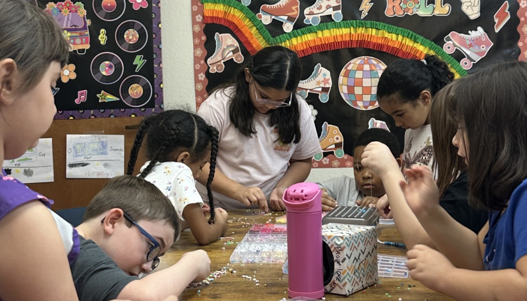 Diverse group of young students sitting around a table, crafting bead bracelets in a classroom.