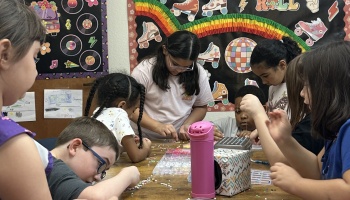 Diverse group of young students sitting around a table, crafting bead bracelets in a classroom.