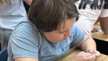 A young student in a blue t-shirt looks down intently while working at a classroom desk.