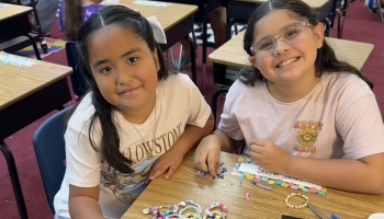 Two young girls smiling at a classroom desk while making colorful beaded bracelets.