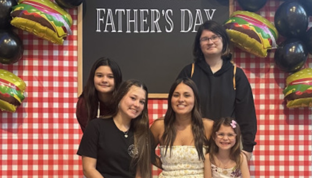 Five smiling girls and young women pose in front of a Father's Day backdrop with hamburger balloons.
