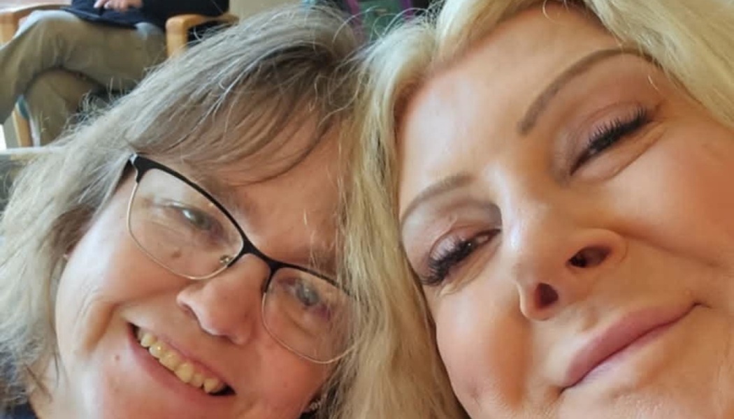 Close-up selfie of two smiling women leaning head-to-head, one wearing glasses and the other with blonde hair.