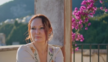 Woman with facial piercings and an embroidered top looking thoughtfully on a balcony with vibrant pink flowers.