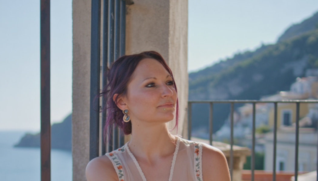 Woman with purple hair smiling gently on a balcony overlooking a sunny coastal town.