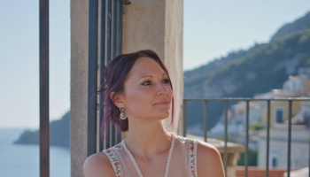 Woman with purple hair smiling gently on a balcony overlooking a sunny coastal town.