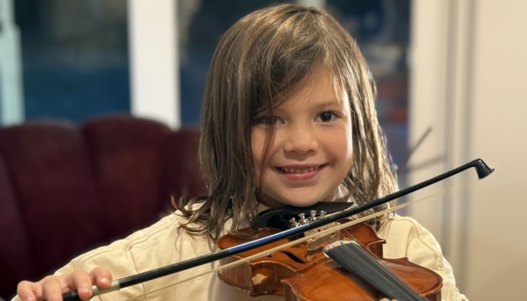 A smiling young child with long hair holds a small violin under their chin with the bow across the strings.