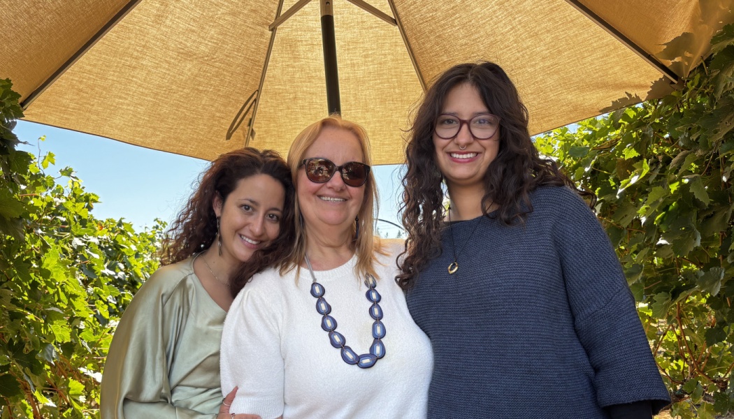 Three women smiling together under a patio umbrella at a vineyard.