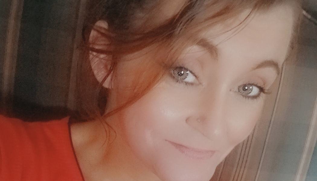 Close-up selfie of a smiling woman with auburn hair, light grey eyes, and a nose stud wearing a red shirt.