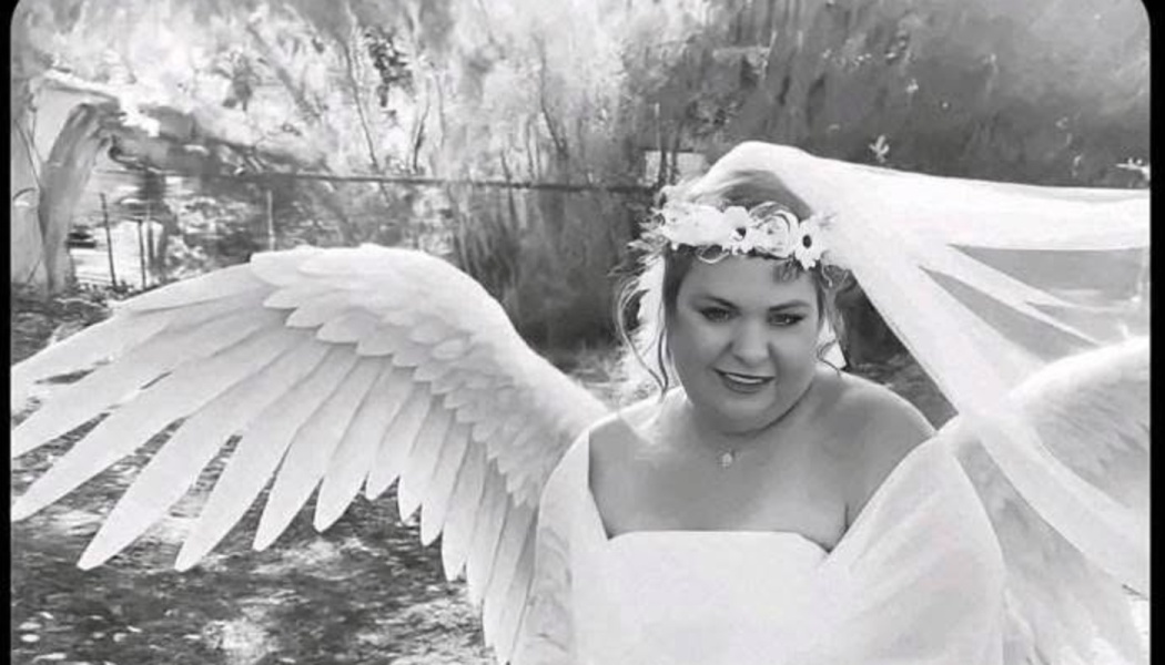 Black-and-white portrait of a smiling woman wearing a white dress, flower crown, veil, and large angel wings.
