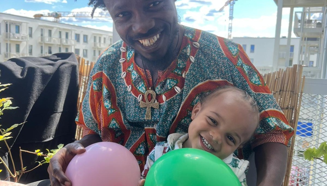 A smiling man in a patterned dashiki holds a cheerful young child, both holding balloons outdoors on a balcony.