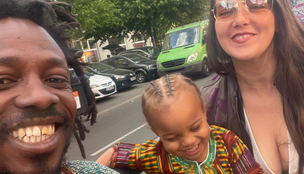 A smiling man with dreadlocks, a woman in glasses, and a happy toddler pose for a selfie on a city street.