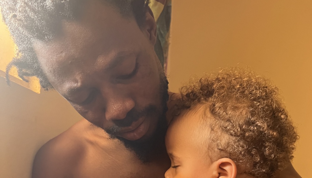 A Black father tenderly holding his sleeping curly-haired child close in a quiet, loving moment.
