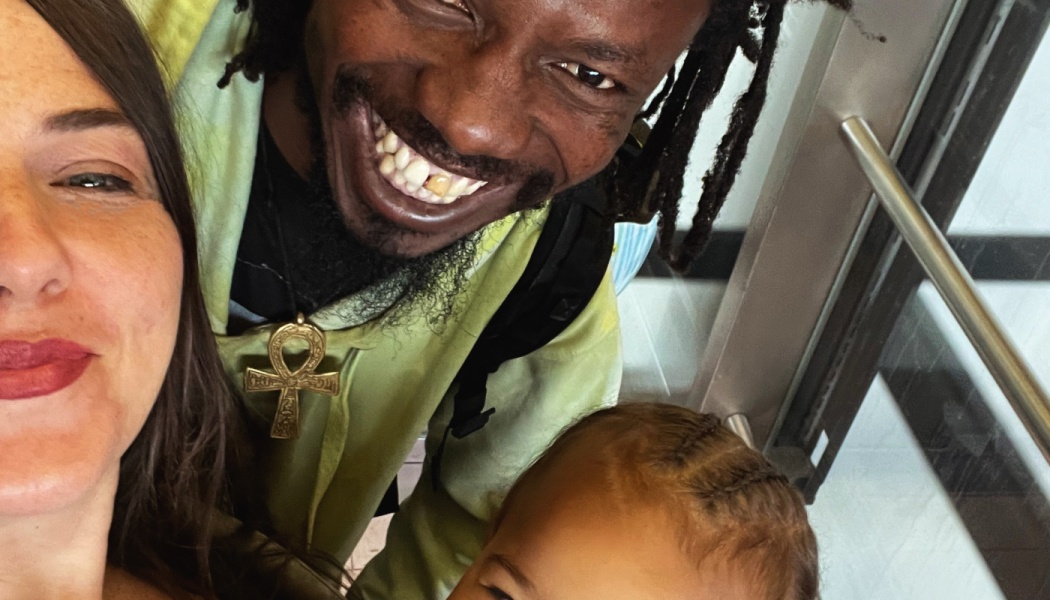 Close selfie of a smiling family: a woman, a cheerful man with dreadlocks and an Ankh necklace, and a young child.