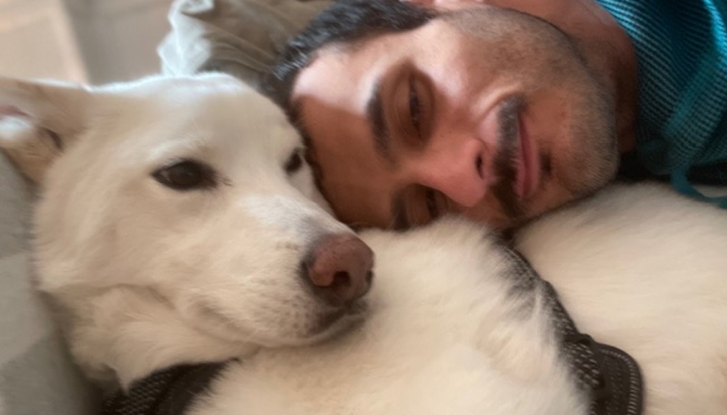 Close-up of a man with a mustache resting his head against a fluffy white dog lying beside him.