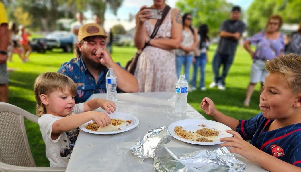 Two young boys eat tacos at an outdoor picnic table while a man laughs in a grassy park.