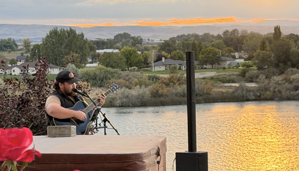 A musician plays acoustic guitar and sings outdoors overlooking a river during a golden sunset.