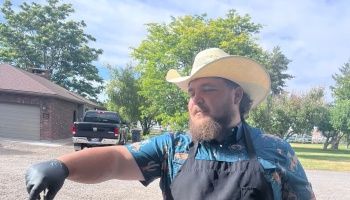 A bearded man wearing a cowboy hat, blue patterned shirt, and black apron standing outdoors.