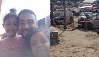 A smiling man taking a selfie with two young girls alongside a crowded tent camp.