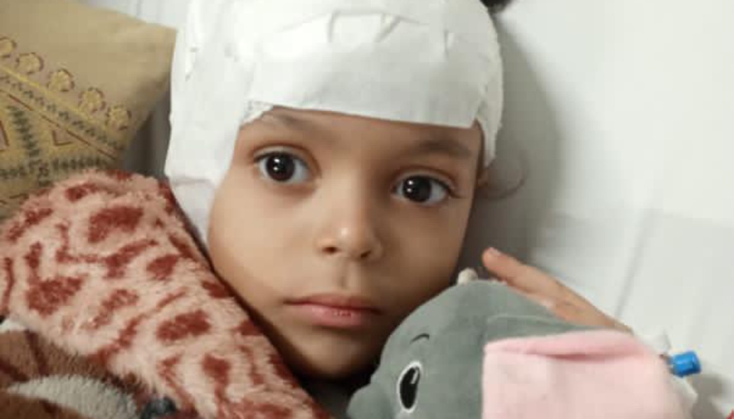 A young child with a white head bandage rests in bed, holding a grey stuffed elephant.