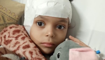 A young child with a white head bandage rests in bed, holding a grey stuffed elephant.
