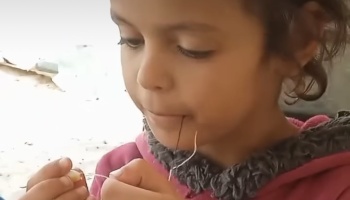 Young girl concentrating as she carefully threads a needle with thread held in her mouth.