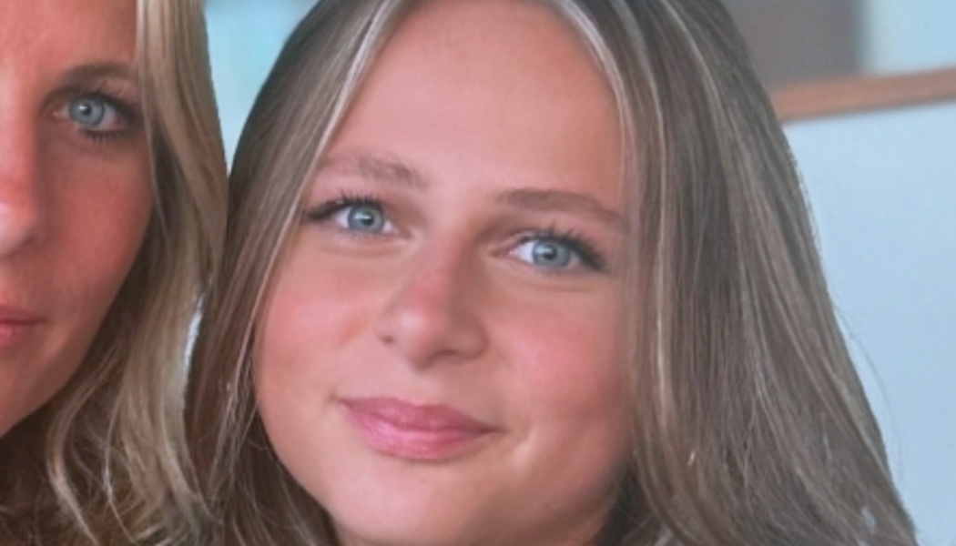 A young blonde woman with blue eyes smiling gently next to another person.
