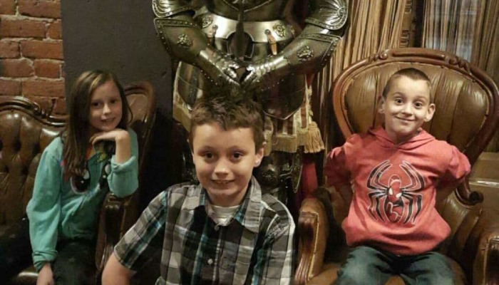 Three smiling children sit in leather armchairs in front of a full suit of shiny armor.