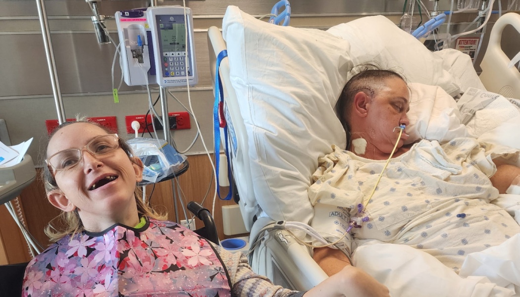 A smiling woman wearing glasses sits beside a patient recovering in a hospital bed with medical equipment nearby.