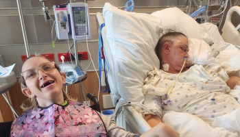 A smiling woman wearing glasses sits beside a patient recovering in a hospital bed with medical equipment nearby.